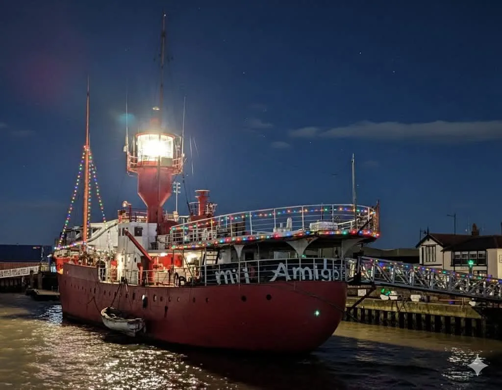 A red ship sits in a dock at night with a quayside to the right. The ship is a light vessel and the lantern is lit.. There is a high mast at the bow and there is a helideck on the sterm; a banner has been hung under the helideck. It reads "Mi Amigo". THis image was AI modified by the originator (originator is known to us)